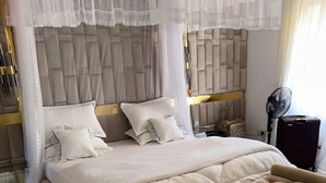 A large bed with a canopy, a bedside table, a fan, and a suitcase. - Island Inn  (Entebbe)