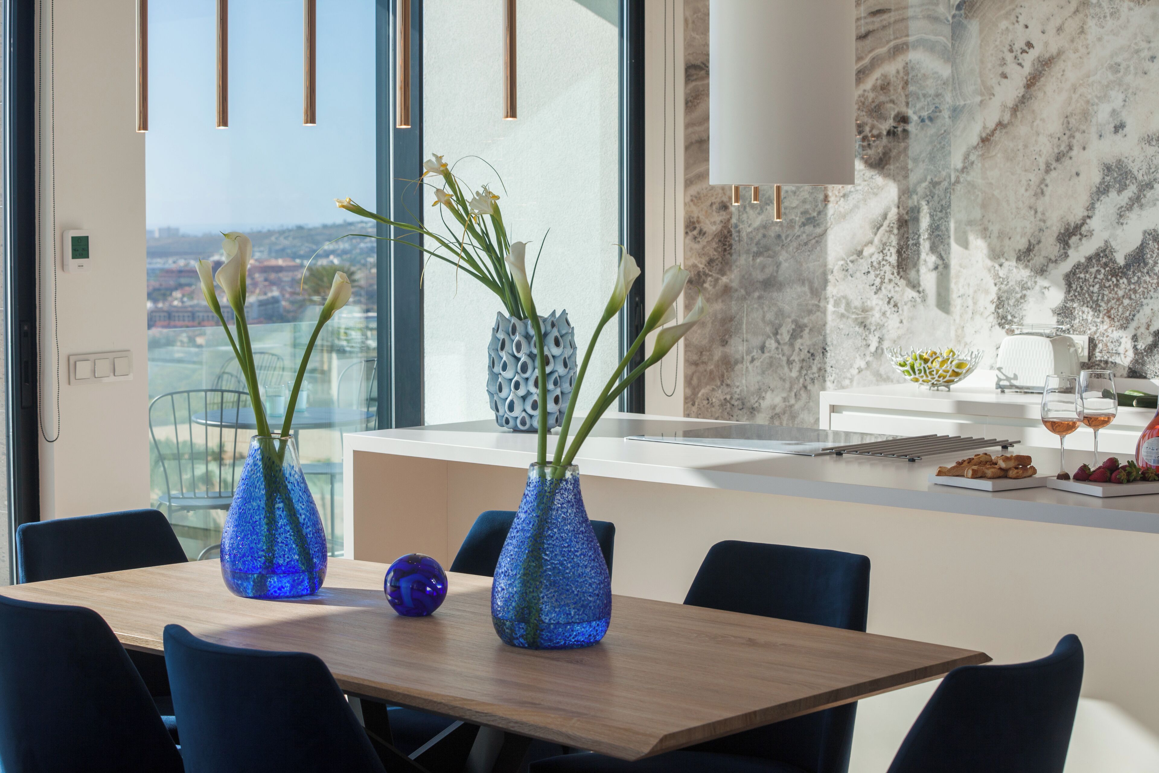 Uma sala de jantar moderna com uma mesa de madeira, dois vasos azuis com flores e vista para um horizonte urbano por grandes janelas.