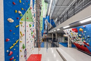 Una struttura indoor di arrampicata con pareti di roccia multiple, un'area imbottita rossa e un piano soppalco con ringhiera.