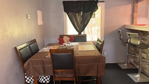 Et spiseområde med et bord dækket til fire personer, stole, et vindue med gardin og et barområde med barstole.