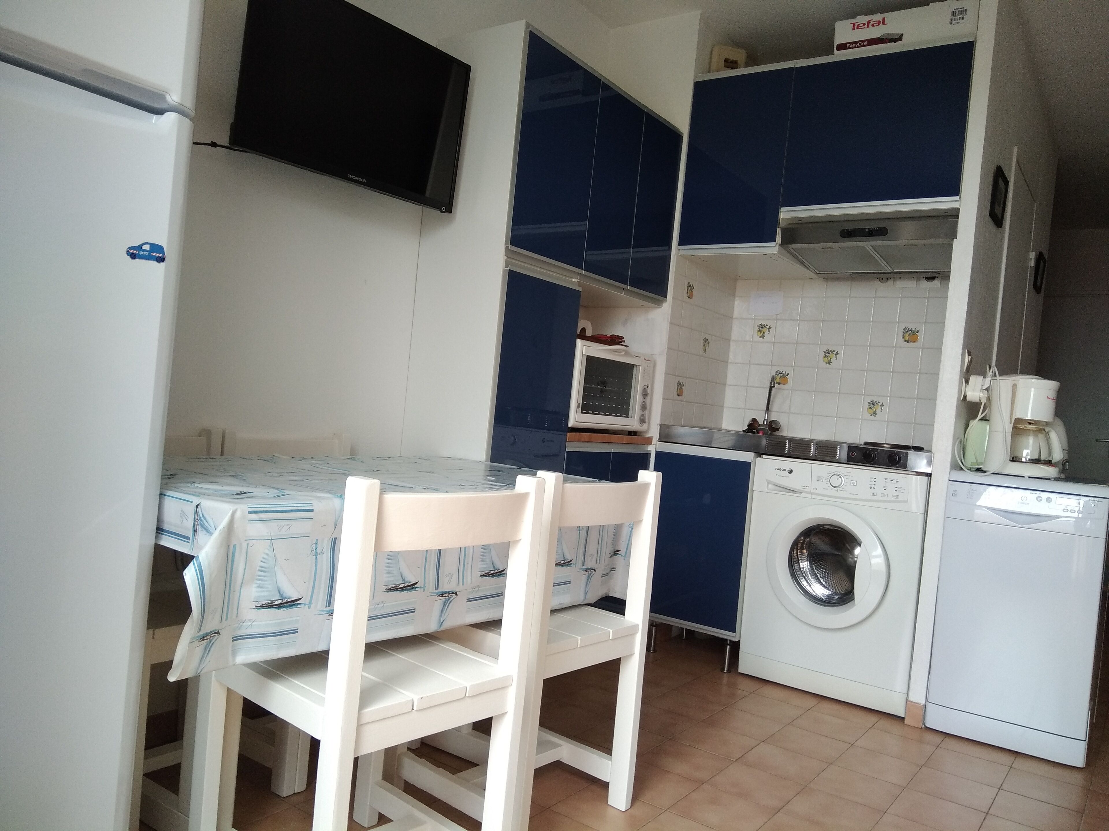Una cocina compacta con una mesa, sillas, lavadora, microondas y un televisor instalado en la pared.