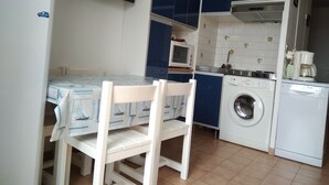 Een kleine keuken met een tafel, stoelen, een wasmachine, een magnetron en een aan de muur bevestigde televisie.