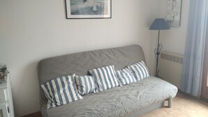 Slaapkamer met een grijze bank, gestreepte kussens, een ingelijst schilderij aan de muur, een vloerlamp en een raam met gordijnen.
