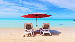 Deux chaises de plage sous un parasol rouge, sur une plage de sable fin, avec une eau turquoise et limpide et un bateau qui se trouve au loin.