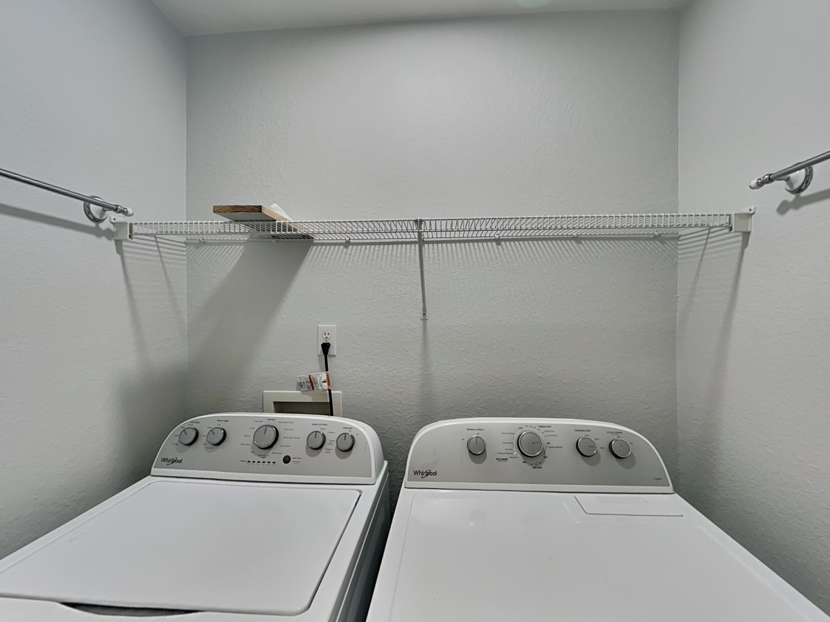 A laundry room with two Whirlpool washers, a metal shelf above, and a power outlet on the wall.