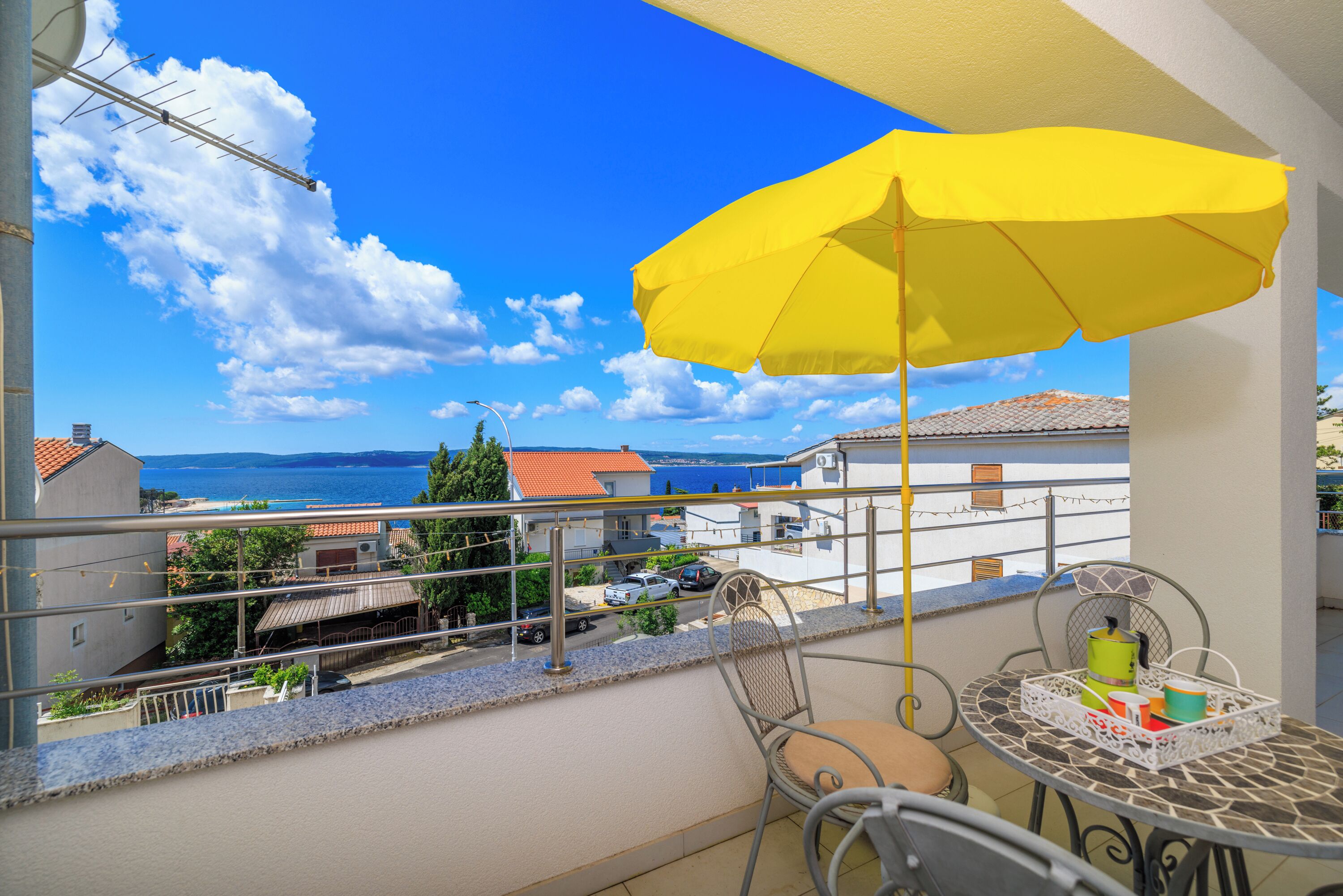 Ein Balkon mit gelber Sonnenschirm, einem Tisch und Stühlen, und Blick auf das Meer und Gebäude.