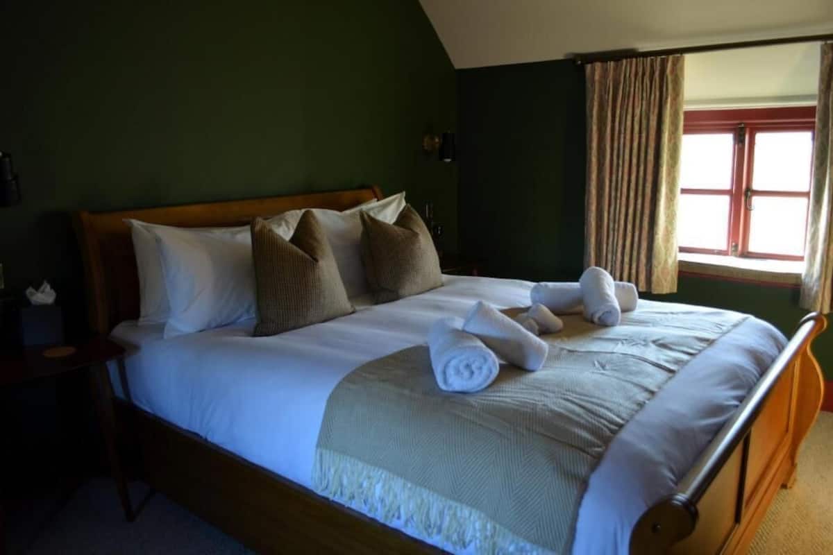 A neatly made bed with pillows and towels, a wooden headboard, a nightstand with a lamp, and a window with curtains.
