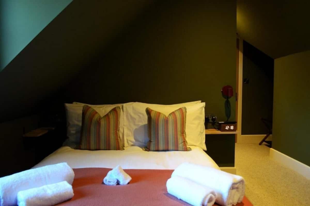 A hotel room with a bed, pillows, a clock, and a vase.