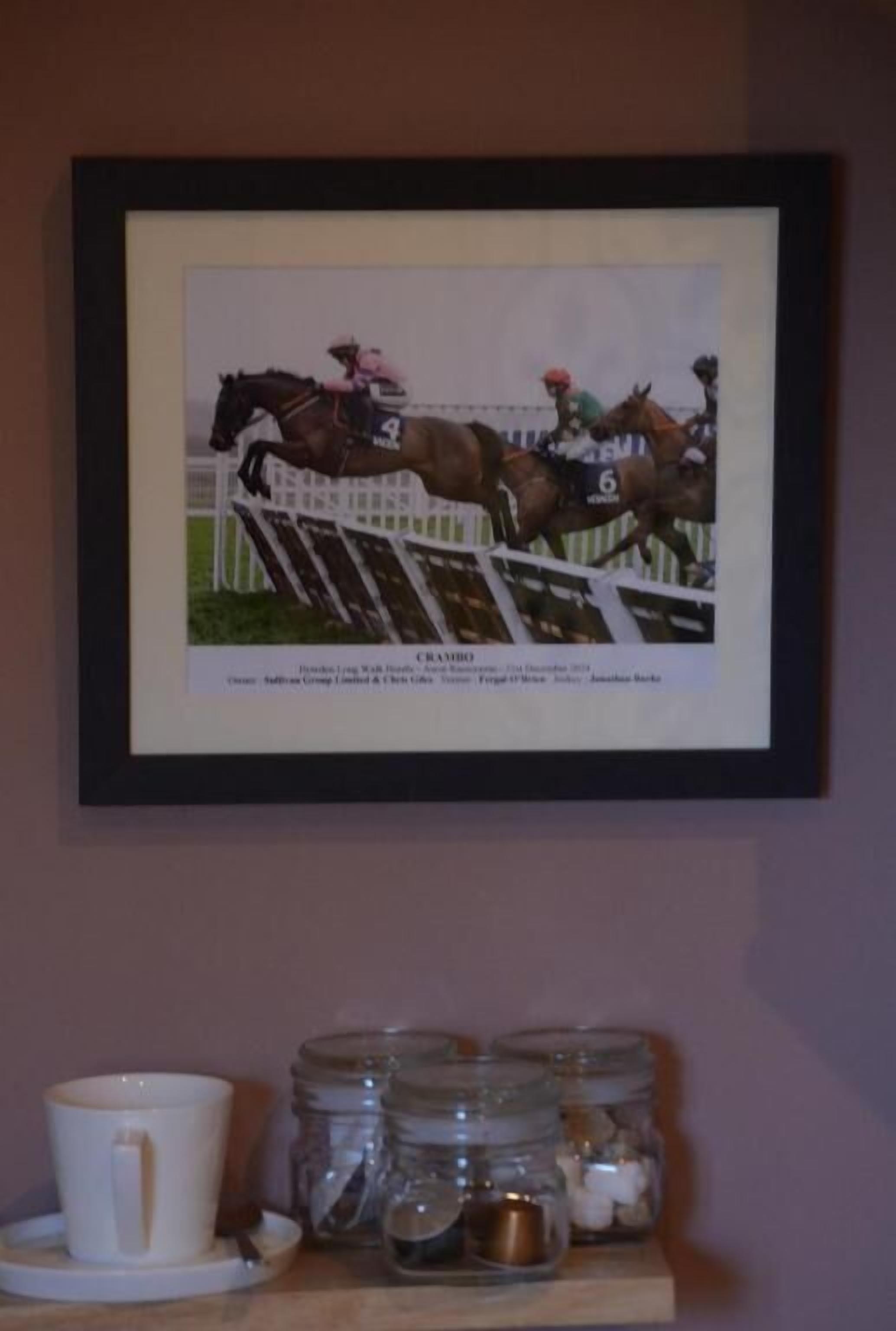 Ein gerahmtes Foto von einem Pferderennen mit einem Pferd mit der Nummer 4 und 6, einem Jockey in Rosa und einem Reiter in Grün. Darunter befinden sich eine weiße Tasse, ein Untertasse und mehrere Gläser mit Deckeln.