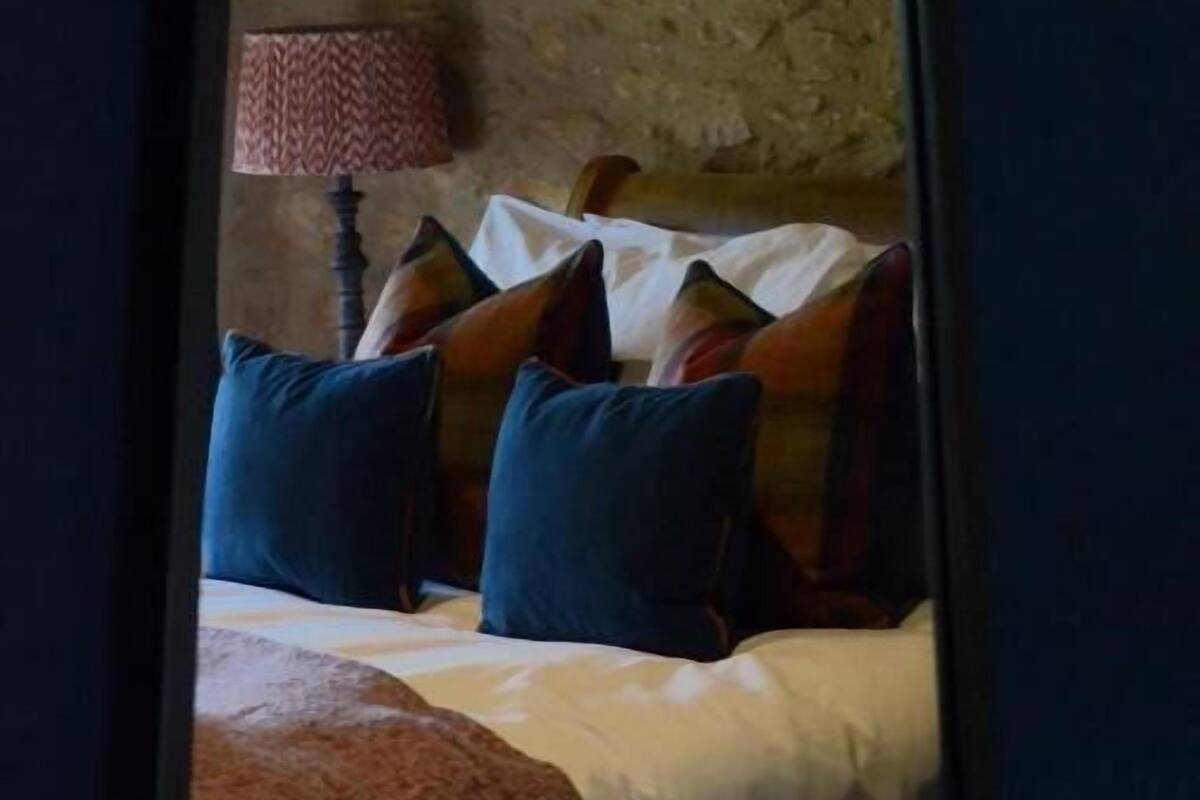 A bedroom with a stone wall, a bed with blue and patterned pillows, and a red patterned lamp.
