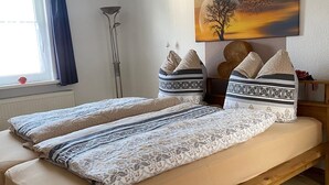 A bedroom with a wooden bed, patterned bedding, a window with curtains, a floor lamp, and a painting on the wall. - BergZeit Karles | Winterberg—Your Favorite Spot on the Rothaarsteig•Ski•Hiking•Bike Park (Winterberg)