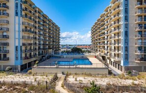 A high-rise residential building with multiple balconies, a central swimming pool, and a sandy foreground. - Watch Sunday fireworks at Northside Park From Your Balcony! (Ocean City)