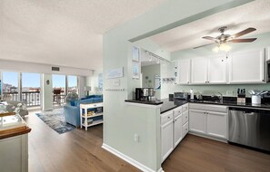 A modern kitchen with white cabinets, a black countertop, and stainless steel appliances. There is a ceiling fan, a dining area with a blue sofa, and a view through large windows. - Watch Sunday fireworks at Northside Park From Your Balcony! (Ocean City)
