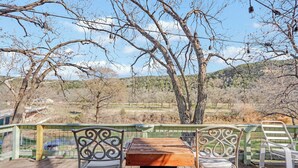 A wooden deck with metal chairs and a table, overlooking a natural landscape with trees and hills. - Guadalupe Riverfront Home | Decks, Firepit & Private River Access (Canyon Lake)