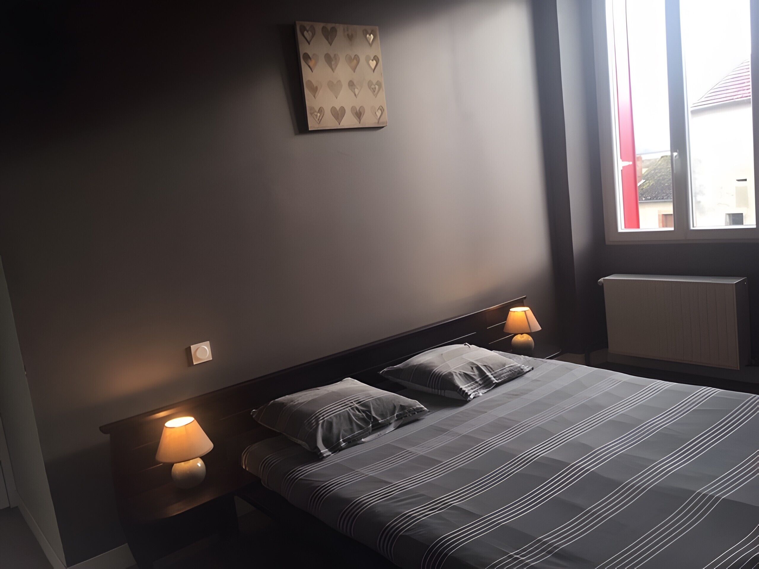 A bedroom with a bed, two bedside lamps, a radiator, and a window.