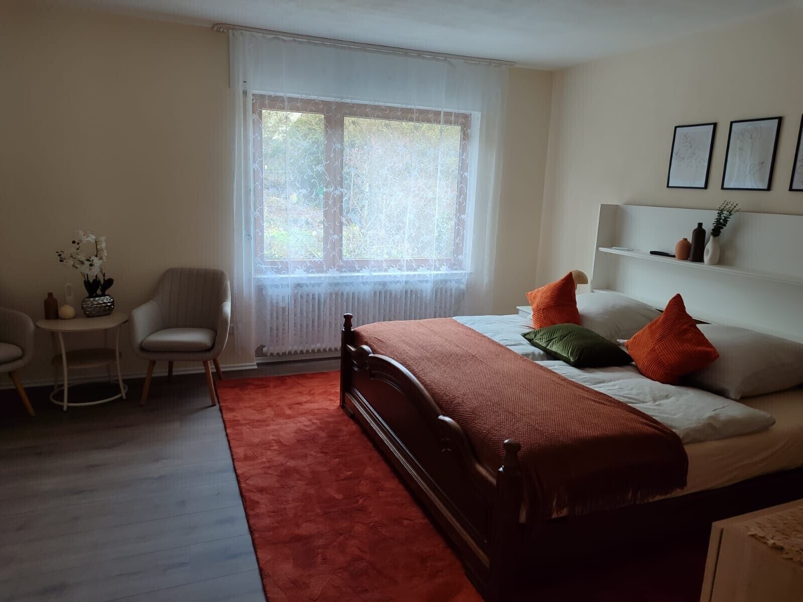 A bedroom with a bed, a chair, a small table, a window with curtains, and a shelf with decorative items.