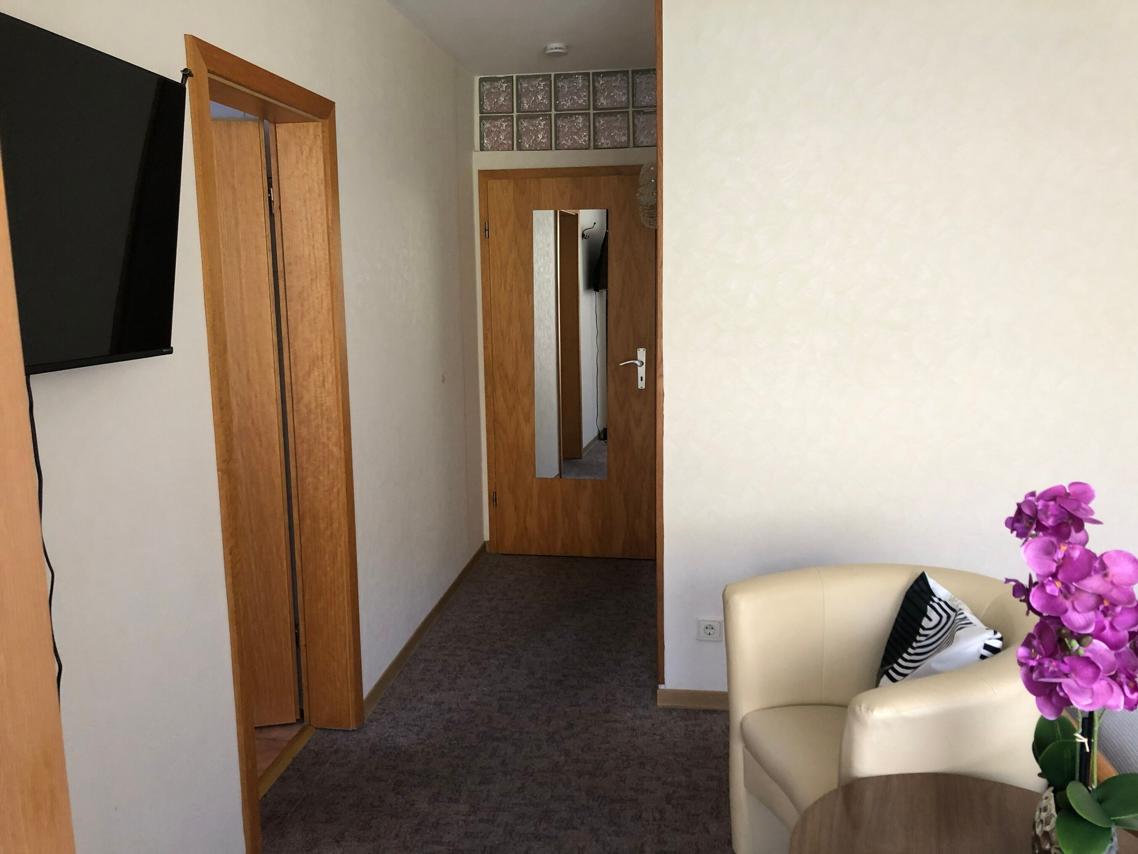 Un couloir avec une porte en bois, un miroir, un téléviseur fixé au mur, un canapé beige orné d’une orchidée violette et un coussin à motifs.