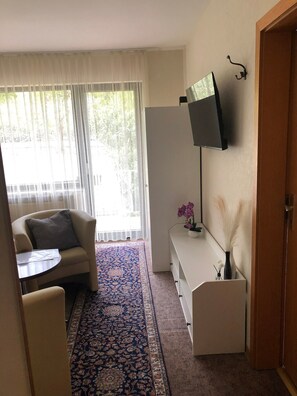 A living room with a sofa, a small table, a flat-screen TV mounted on the wall, a decorative vase, and a patterned rug. - Room 'Zimmer 1 OG' with Shared Garden, Balcony and Wi-Fi (Klotten)