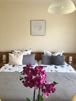 A neatly made bed with floral bedding, two pillows, a framed artwork on the wall, and a vase with purple flowers in the foreground. - Room 'Zimmer 1 OG' with Shared Garden, Balcony and Wi-Fi (Klotten)