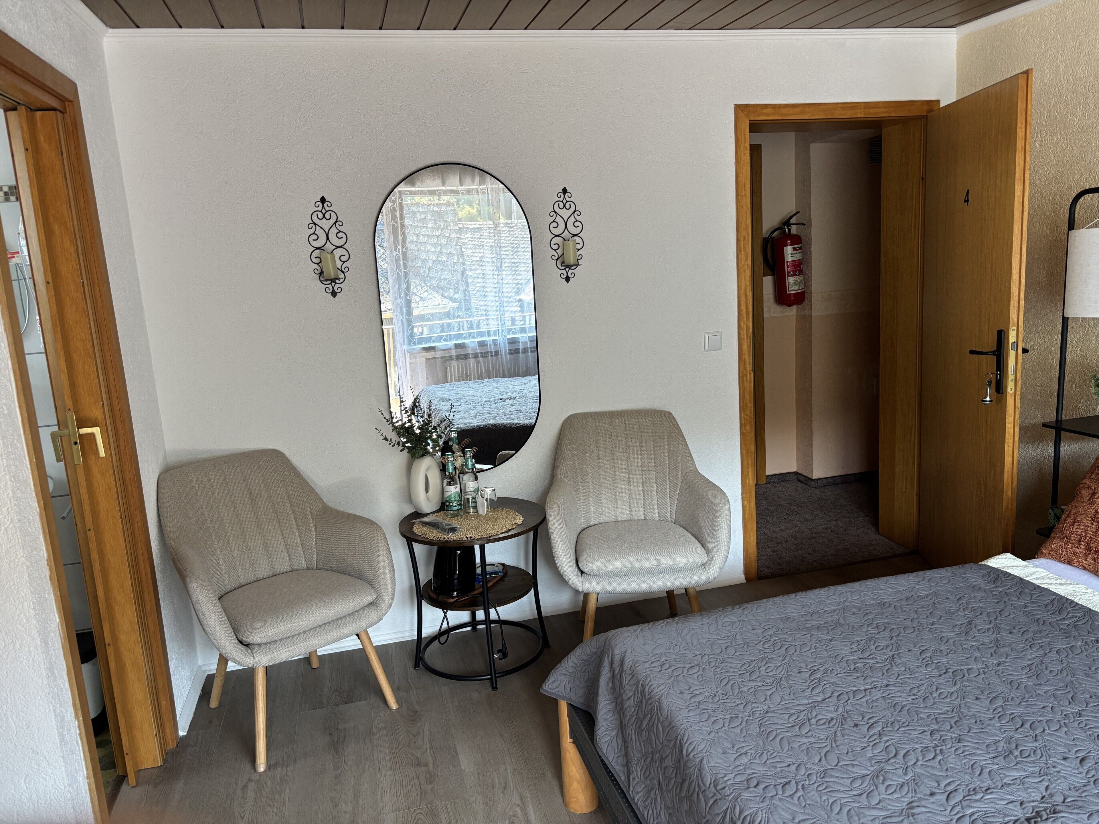Ein Hotelzimmer mit einem Bett, zwei Sesseln, einem kleinen Tisch mit einer Vase und Flaschen, einem Spiegel und einer Tür zu einem weiteren Raum.