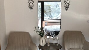 Ein Zimmer mit zwei Sesseln, einem kleinen runden Tisch mit einer Vase und Weingläsern sowie einem großen Spiegel an der Wand.