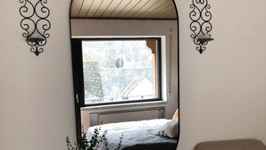 Ein Zimmer mit einem großen Spiegel, zwei Sesseln, einem kleinen Tisch mit einer Vase und Weingläsern sowie einem Fenster mit Blick auf Gebäude.