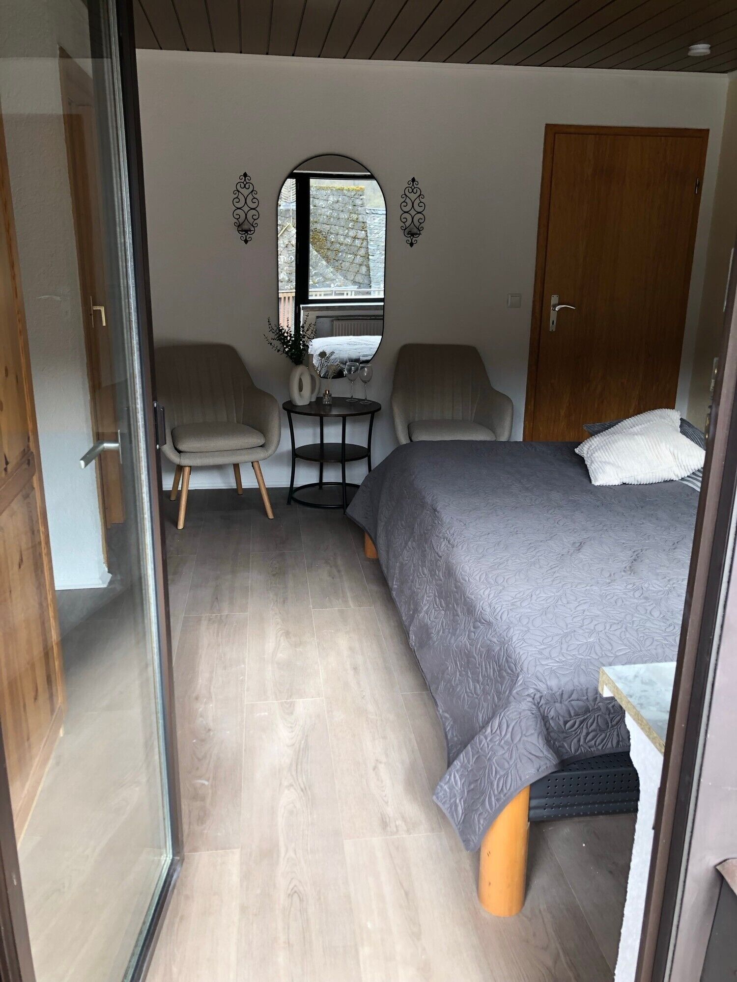 Ein gemütliches Schlafzimmer mit einem Bett, zwei Stühlen, einem kleinen Tisch mit einer Vase und Gläsern, einem Spiegel und einer Holztür.