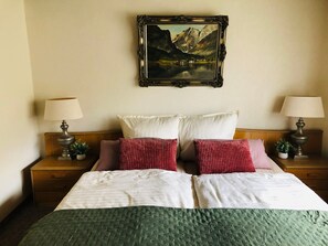 A double bed with a green quilt, two bedside tables with lamps, a framed painting of a mountain landscape, and a wooden headboard. - Room 'Zimmer 3D, Antik 2og' with Shared Garden, Balcony and Wi-Fi (Klotten)