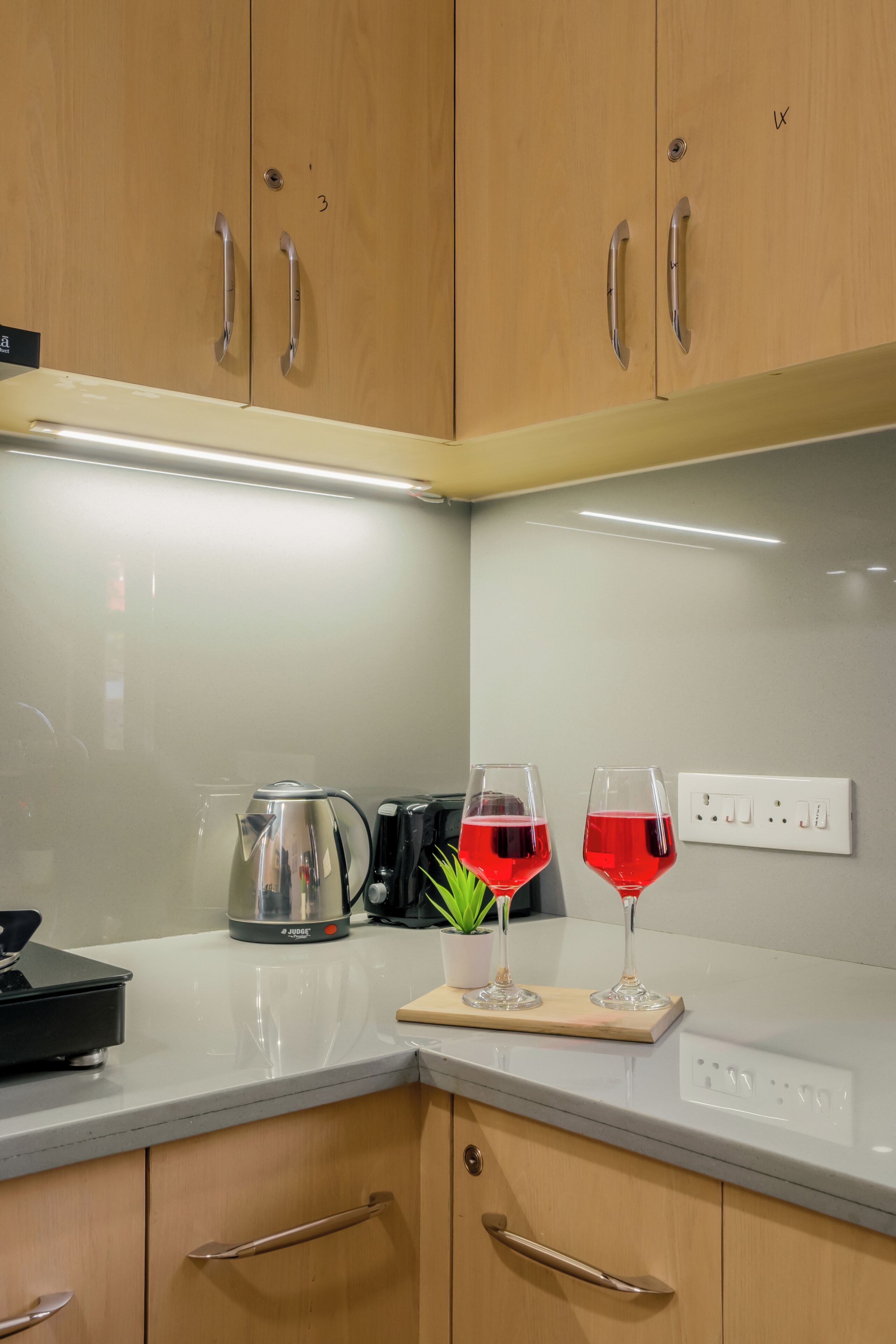 A kitchen with wooden cabinets, a stainless steel kettle, a cutting board with glasses, and a potted plant.