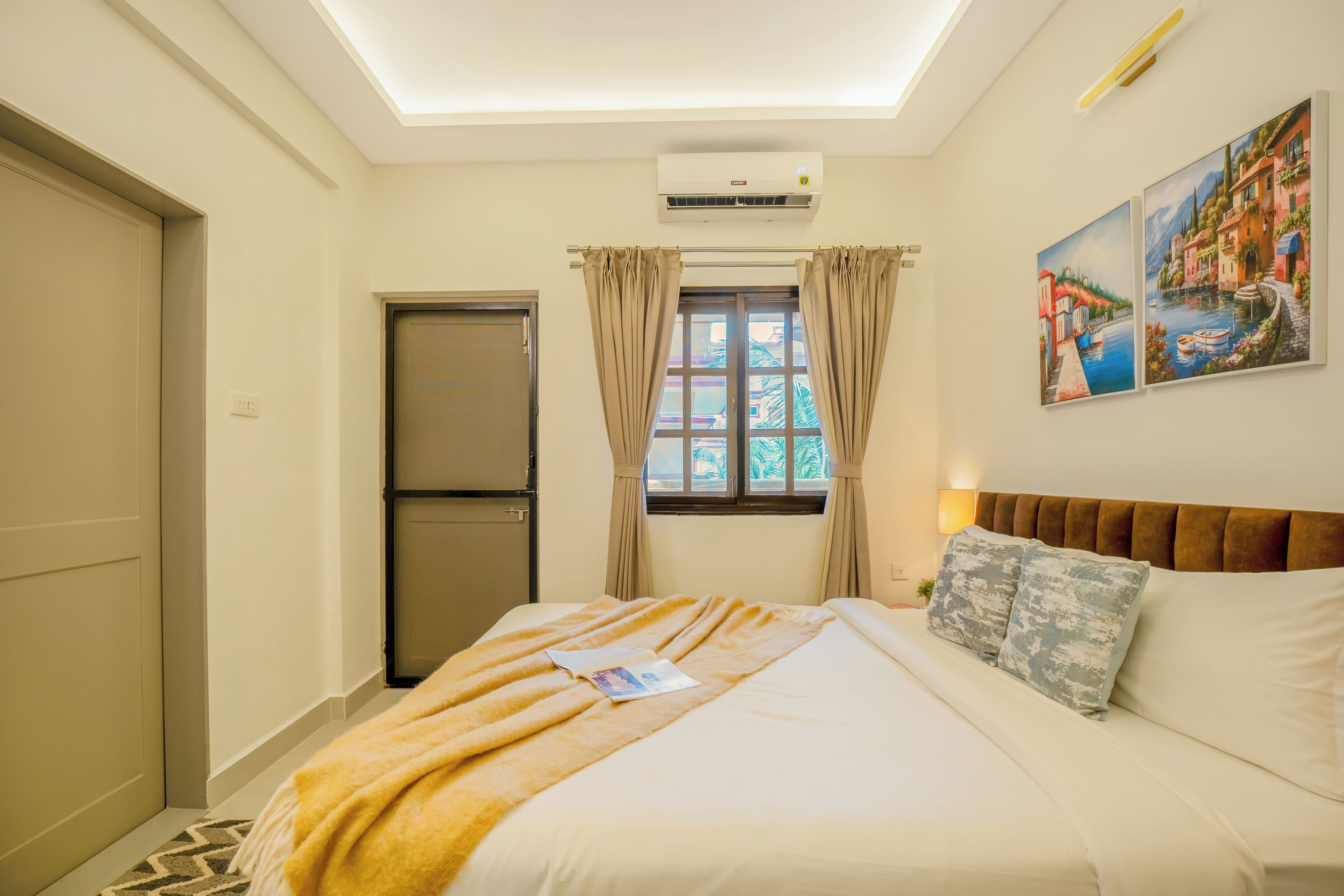 A hotel room with a bed, a window with curtains, an air conditioning unit, and a painting on the wall.