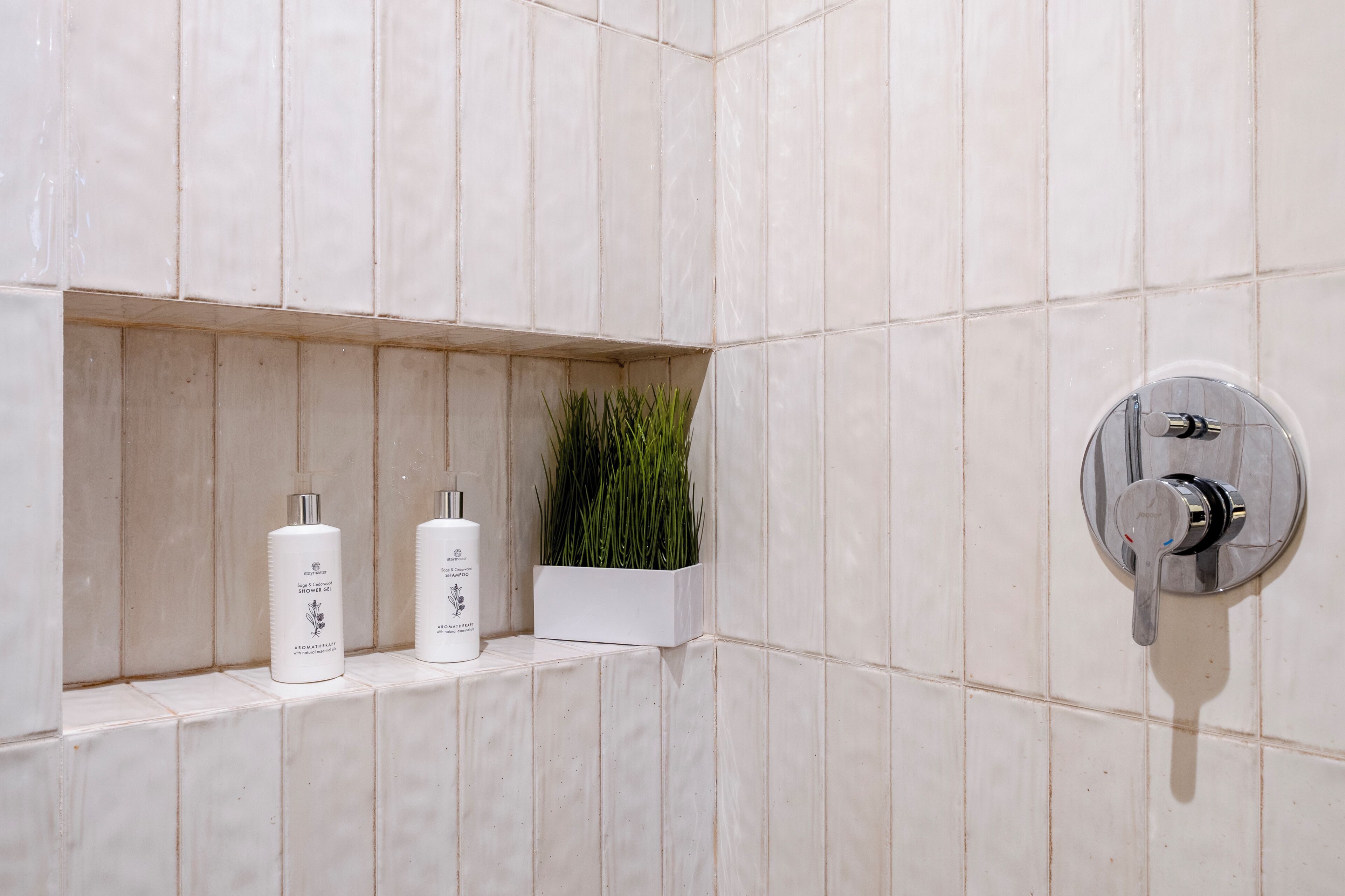 A bathroom with white tiles, a showerhead, and two bottles of shampoo and conditioner on a shelf.