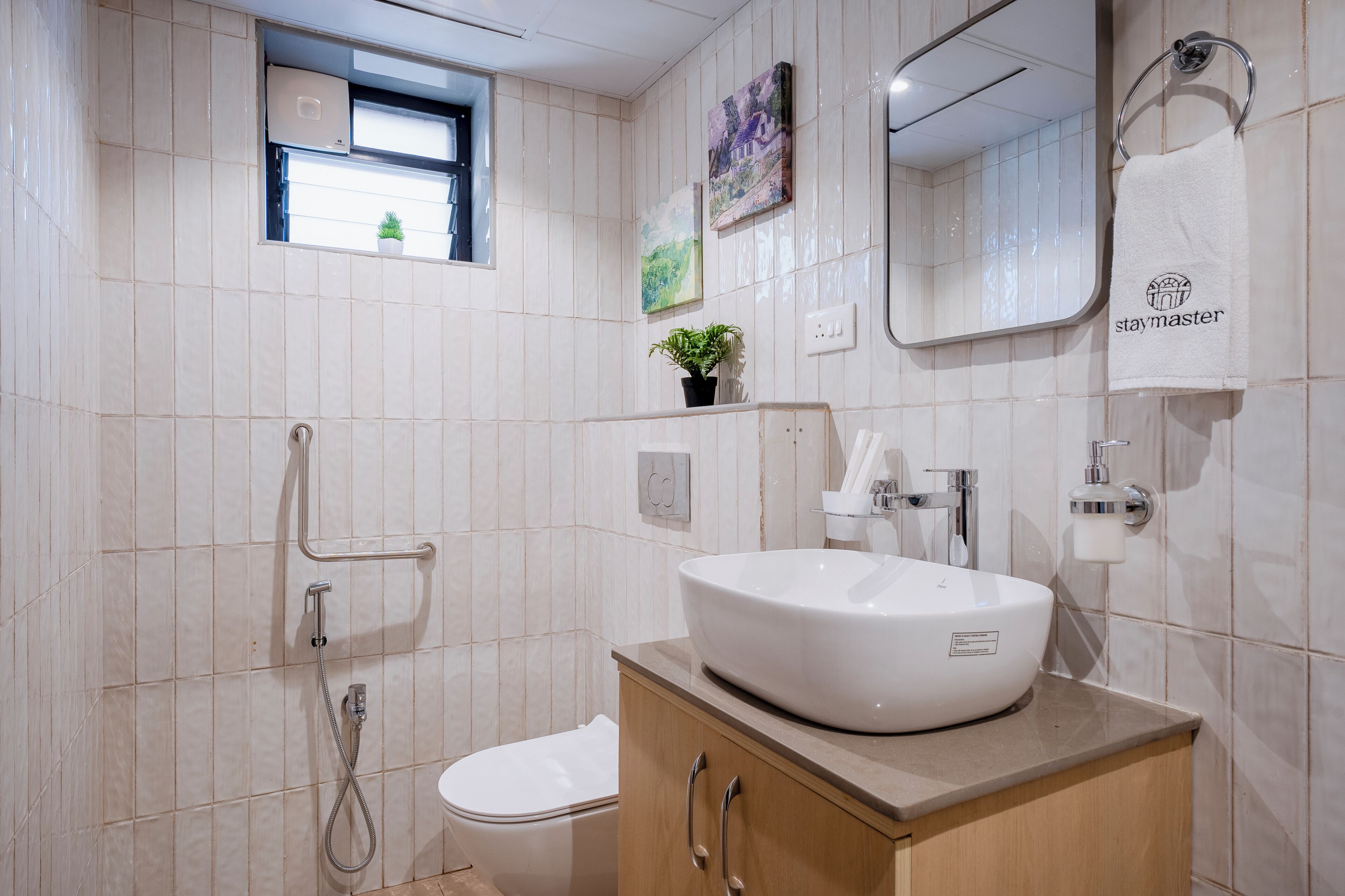 Un baño moderno con lavamanos blanco, inodoro, ducha y una pequeña ventana con una planta.