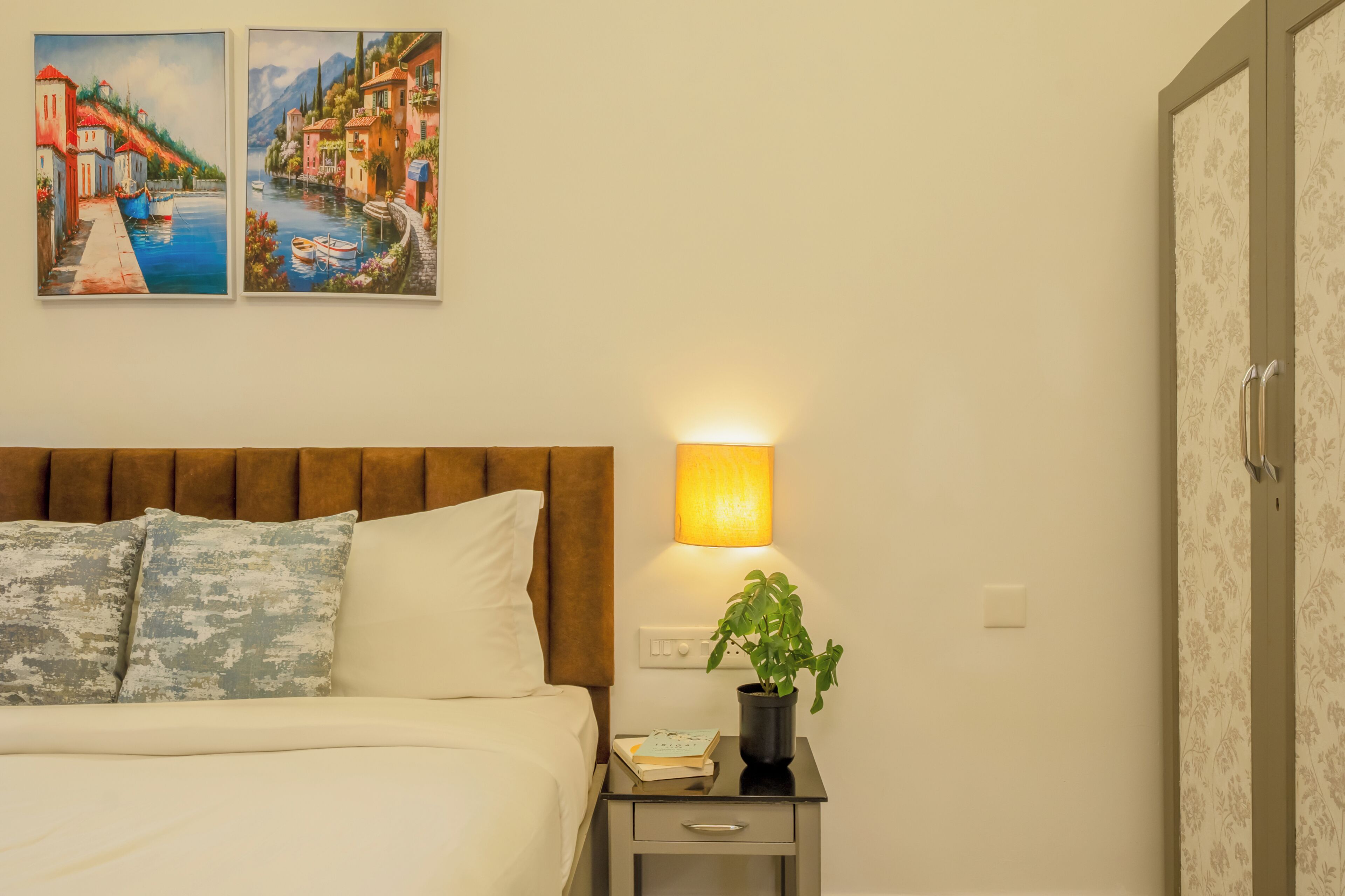 A neatly made bed with pillows, a bedside lamp, a small table with a book and a plant, and two paintings on the wall.