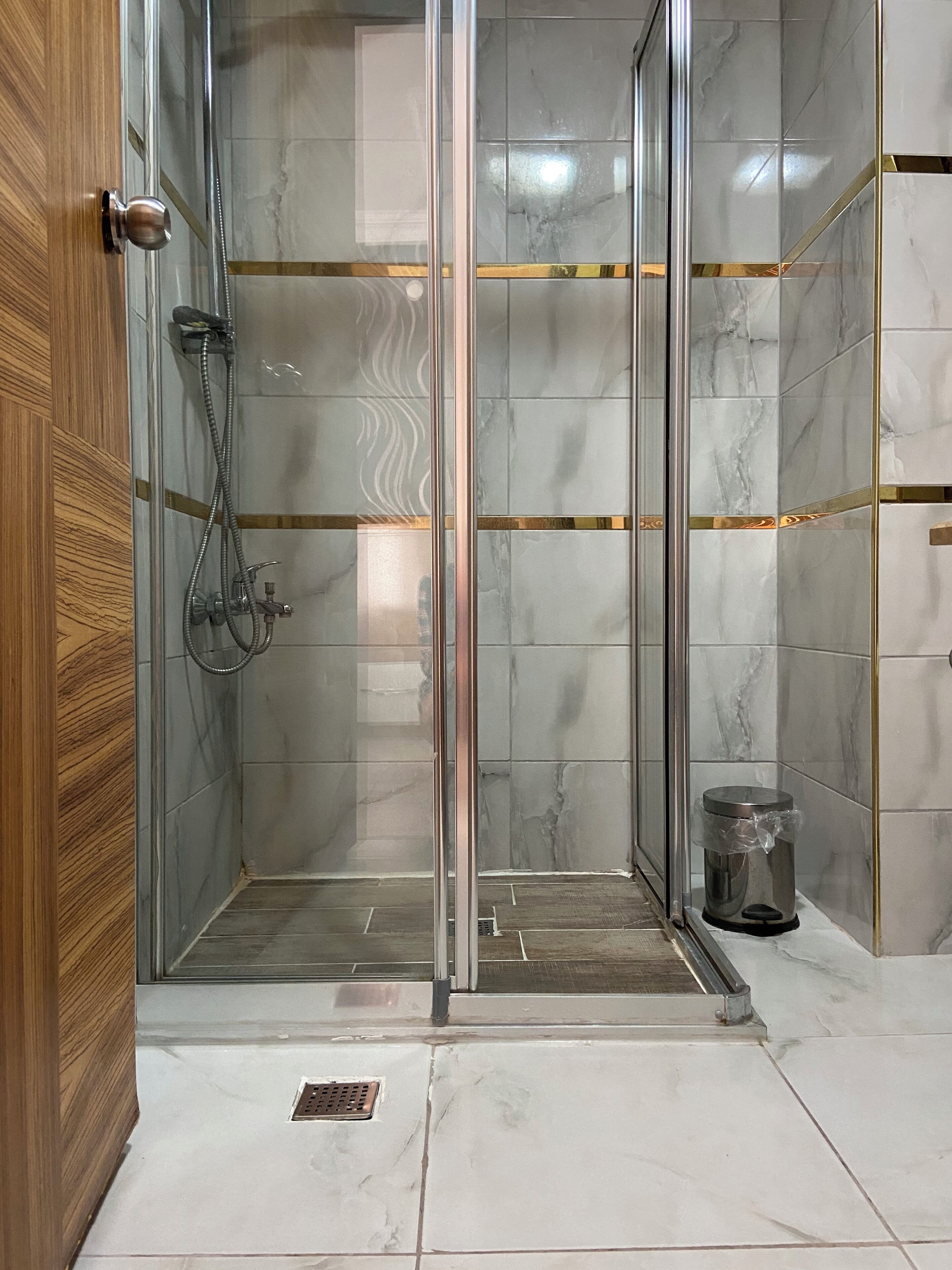 A glass-enclosed shower area with a wooden door, tiled walls, and a trash bin.