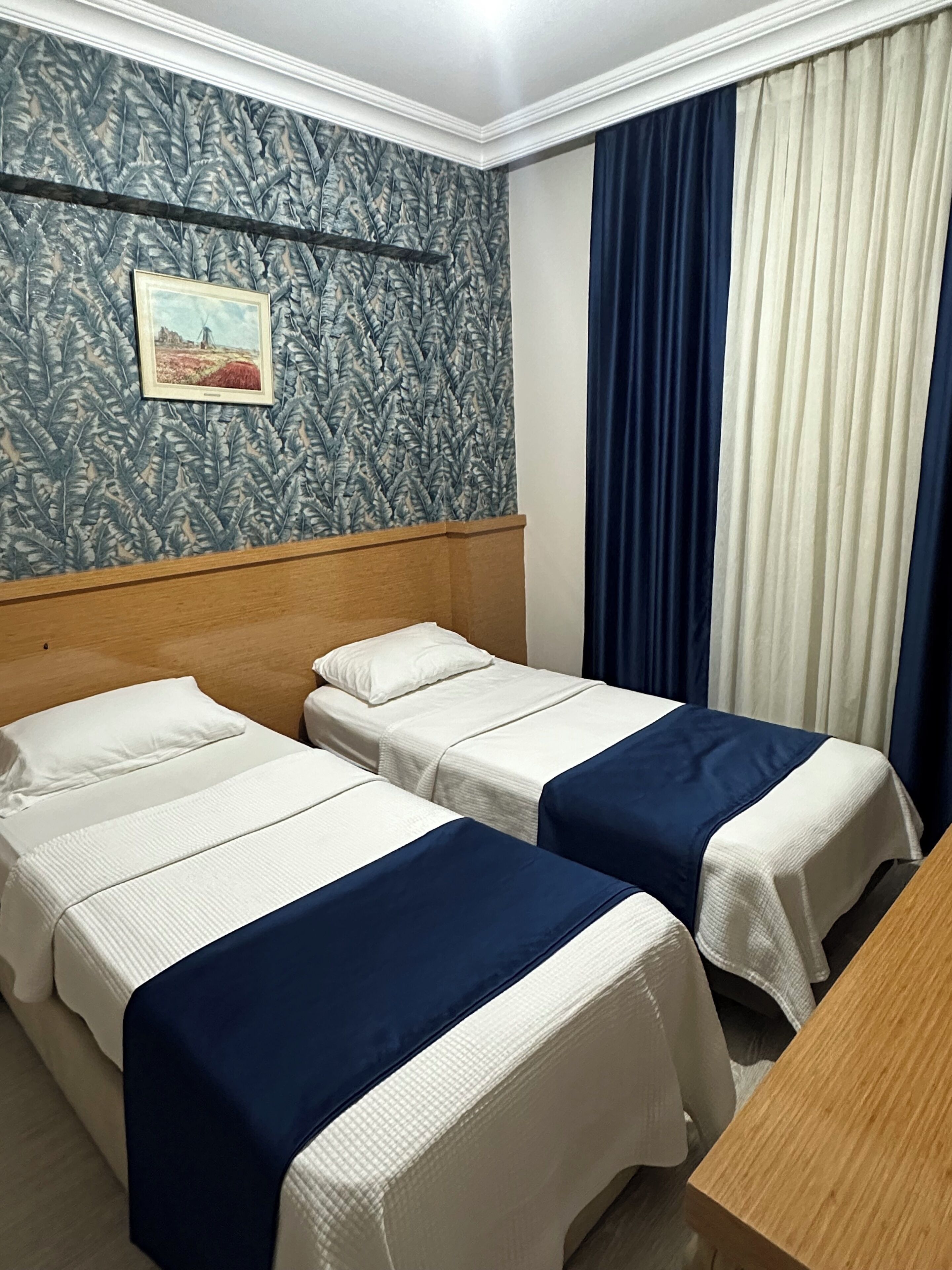 Une chambre d’hôtel avec deux lits, une tête de lit en bois, un papier peint à motifs, un tableau et une fenêtre avec des rideaux.