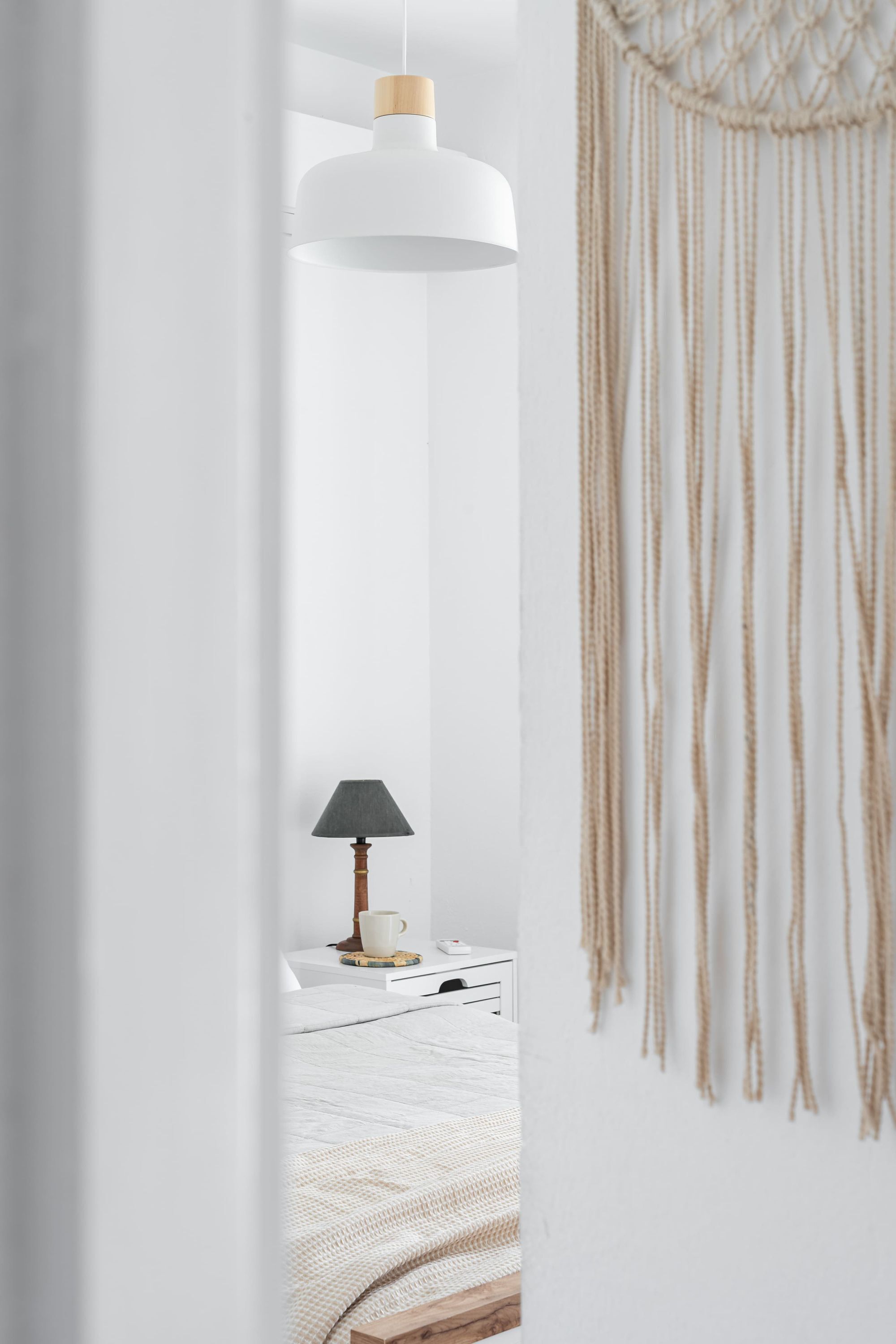 A bedroom with a bed, a bedside table with a lamp, a macrame wall hanging, and a hanging light fixture.