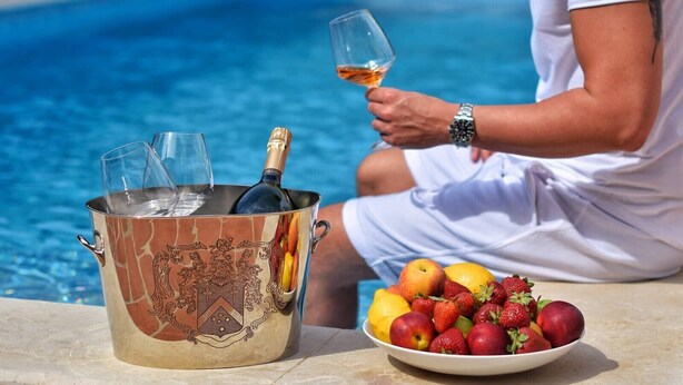En person sidder ved en pool med en champagneflaske og glas i en dekorativ spand, samt en skål med frugt på bordet.