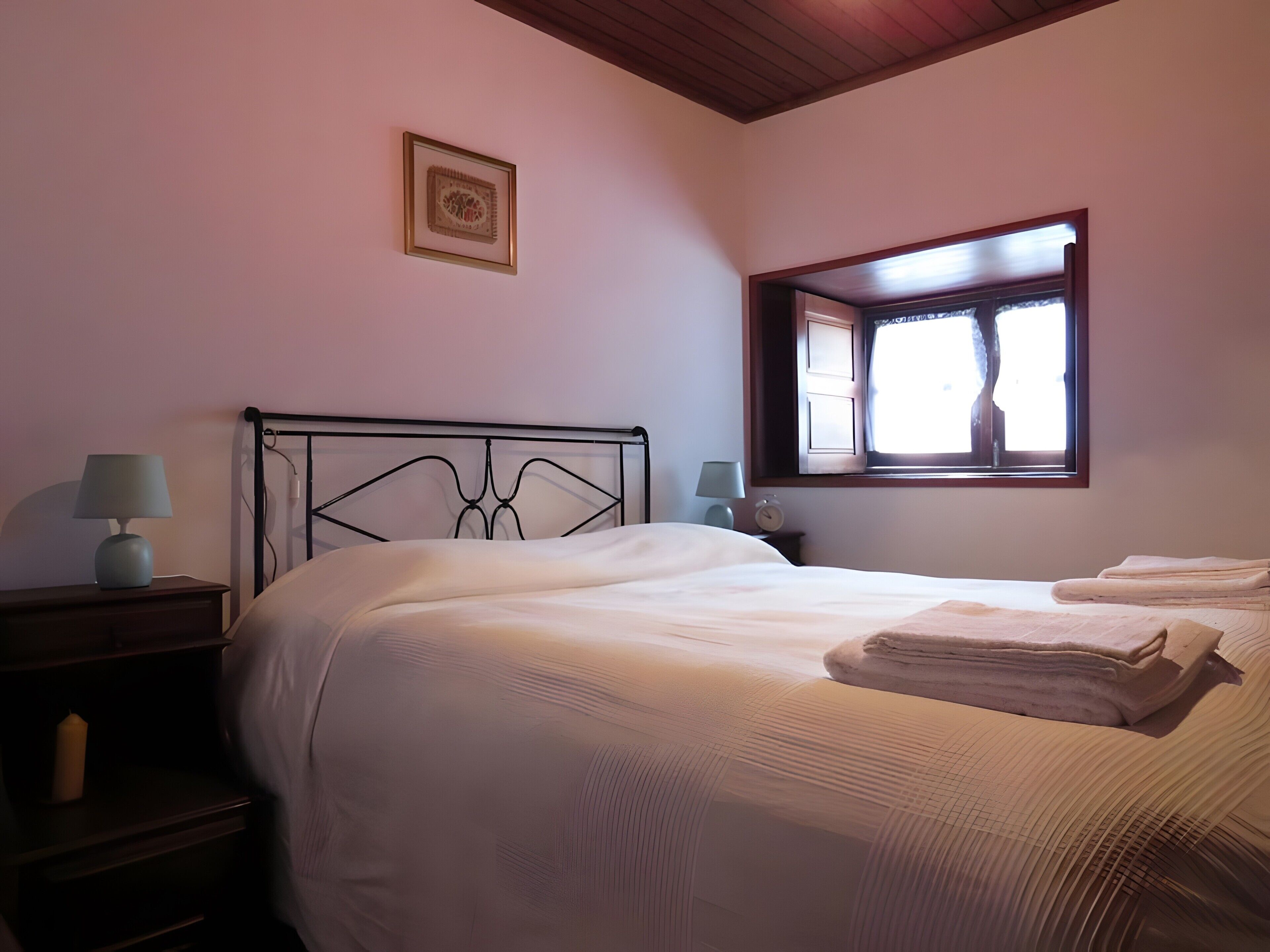Un dormitorio con una cama, dos mesitas de noche con lámparas, una ventana con cortinas y un cuadro en la pared.