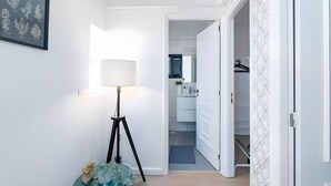 Ein Flur mit Stehlampe, einem gerahmten Pflanzenbild und einer offenen Tür zu einem Badezimmer mit weißem Waschbecken und Spiegel.