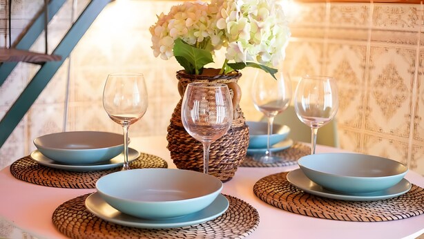 Ein gedeckter Tisch mit blauen Tellern, klaren Weingläsern und einem geflochtenen Korb voller weißer Blumen.