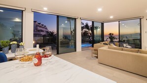 En moderne stue med en stor sofa, et spisebord med en vinflaske, og udsigt til en swimmingpool samt bygninger i skumringen.