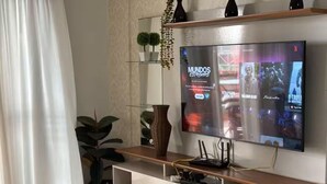 A modern living room with a flat-screen TV, wooden shelves, a wicker chair, and potted plants. - Spacious apartment 160m from the beach, 3 bedrooms, barbecue. (Itapema)