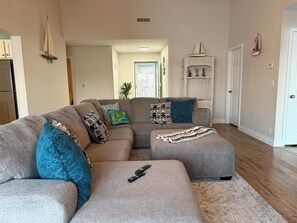 Ein Wohnzimmer mit einem Ecksofa, einem Hocker und einer Fernbedienung auf dem Hocker.