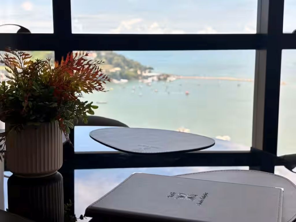 Een tafel met een laptop, een bloempot en uitzicht op de zee door een raam.