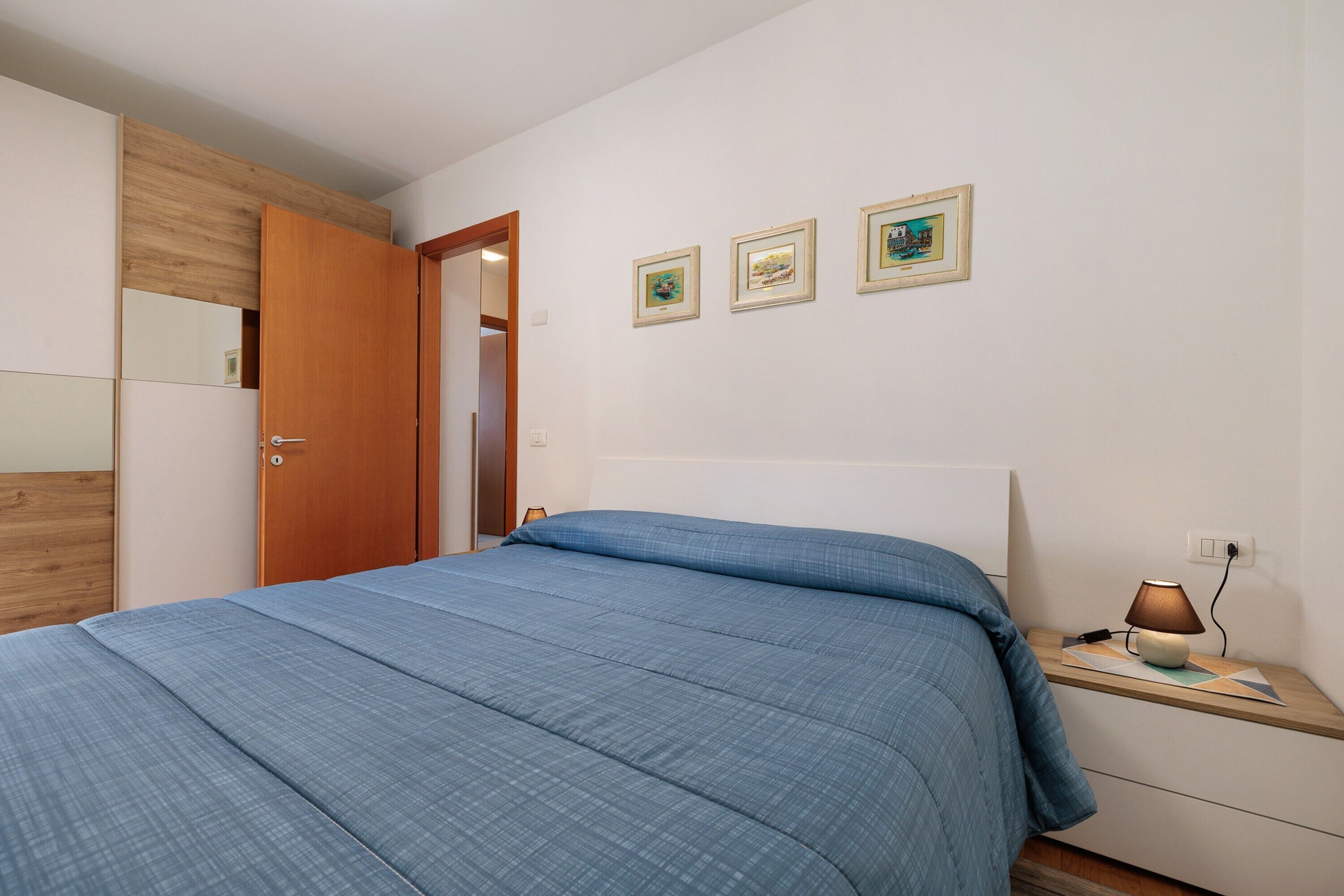Un dormitorio con una cama, una mesita de noche con lámpara, un armario de madera y tres cuadros en la pared.