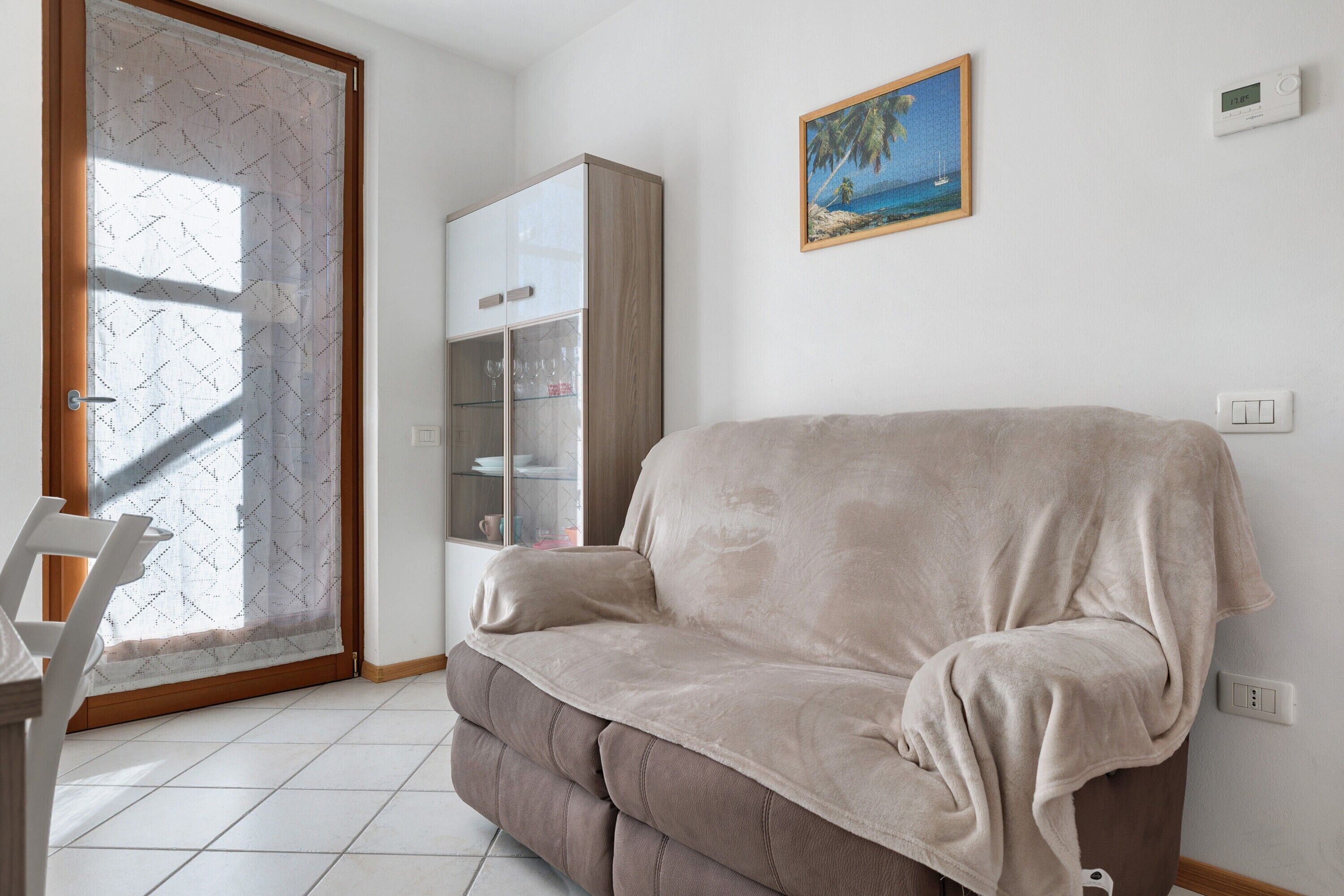 Ein Wohnzimmer mit einem beigefarbenen Sofa, einer Glaskabine, einem Gemälde mit Strandmotiv und einem Thermostat an der Wand.