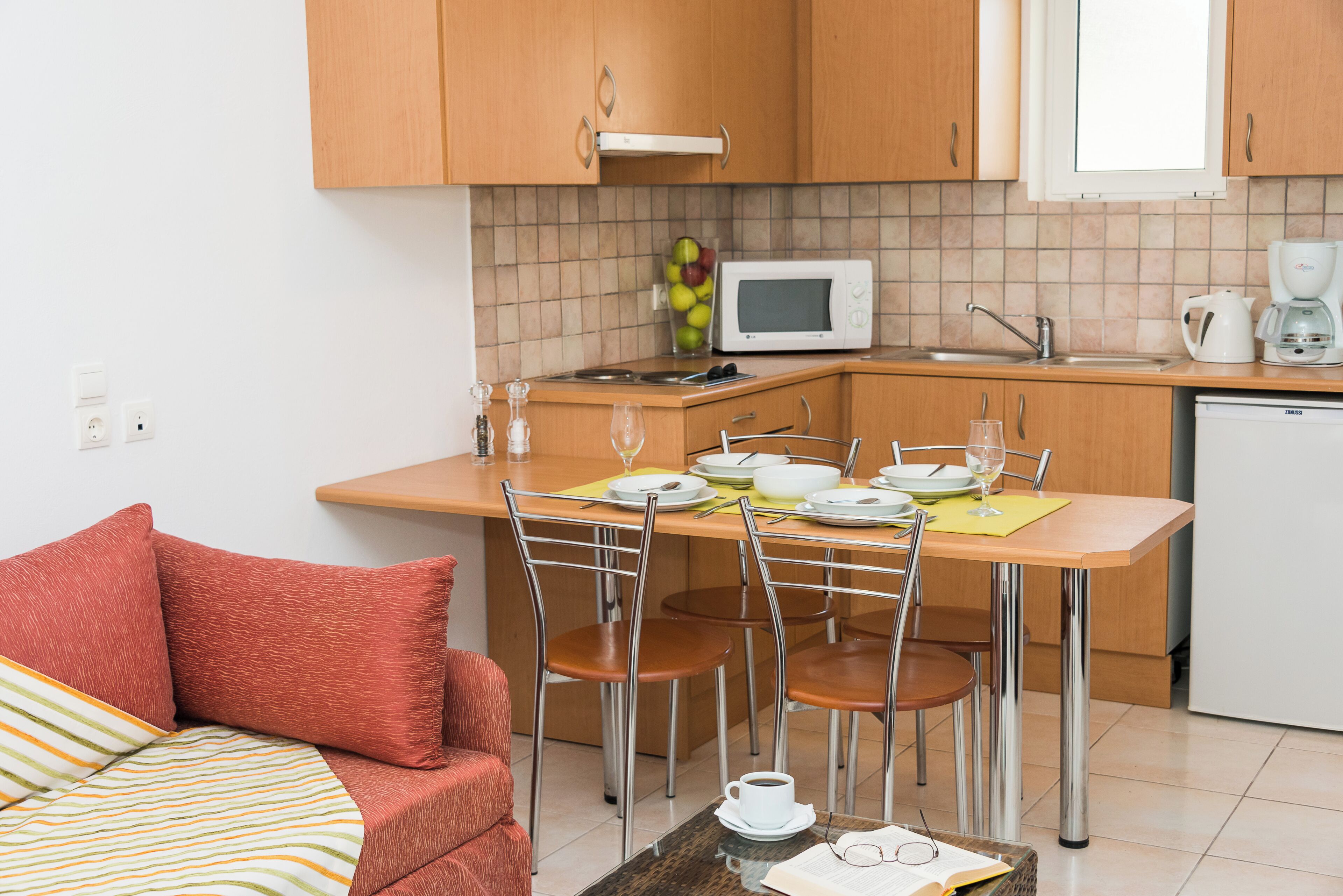 Cocina compacta con microondas, lavaplatos y cafetera. Hay una mesa de comedor con sillas y un pequeño sofá con cojines.