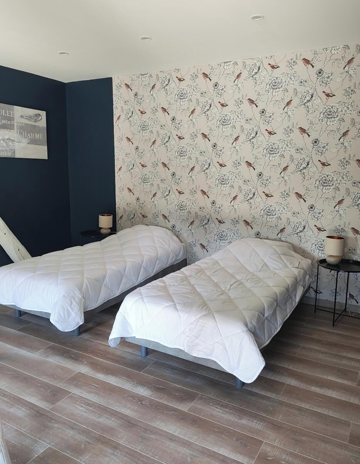 A room with two beds, a dark blue wall, floral wallpaper, and a poster.