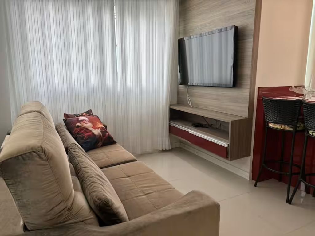 Ein Wohnzimmer mit einer Couch, einem an einer Holzwand montierten Flachbildfernseher, einem roten Tisch mit zwei Stühlen und einem Fenster mit transparenten Vorhängen.