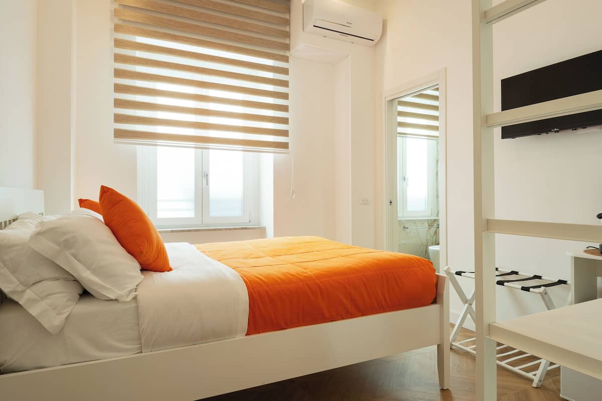 Ein Schlafzimmer mit Bett, weißen Wänden, Holzboden und einem Fenster mit Jalousien.